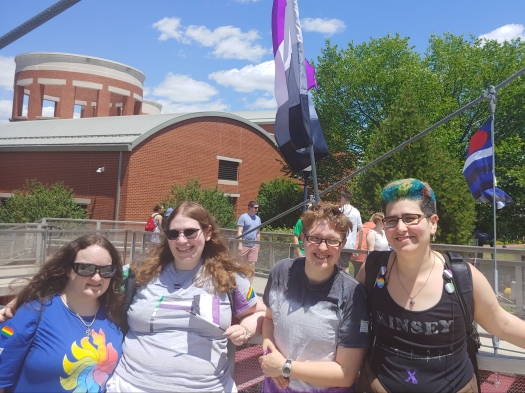 Four people pose outdoors in front of an ace flag. There is a leather flag in the background.
